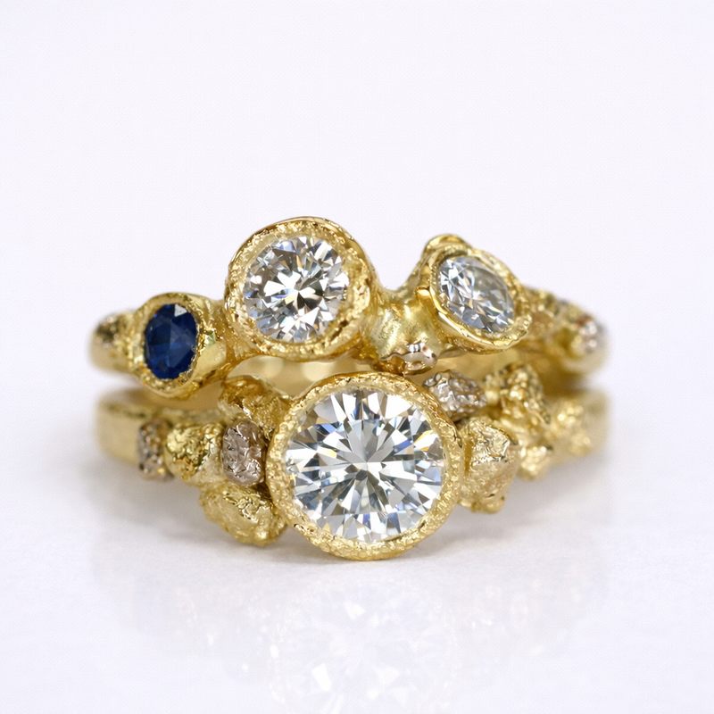Custom organic stacked gold engagement rings featuring round diamonds and a blue sapphire, handcrafted with textured surfaces by Albuquerque jeweler Alexis Pavlantos.