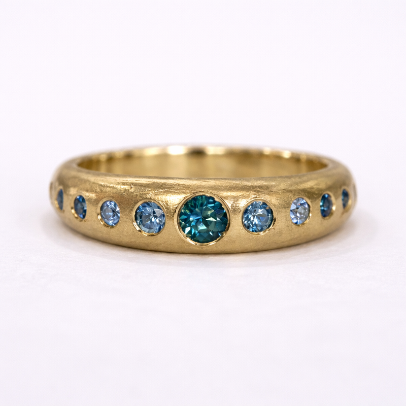 Custom gold band by Alexis Pavlantos set with a row of flush-set blue sapphires, featuring a central teal sapphire surrounded by lighter blue stones.