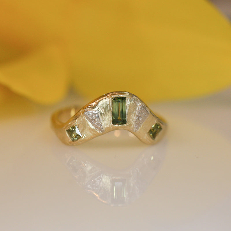 Custom gold contour ring set with an emerald-cut green sapphire and two accent sapphires, handcrafted by Alexis Pavlantos, featuring an organic, sculptural surface.