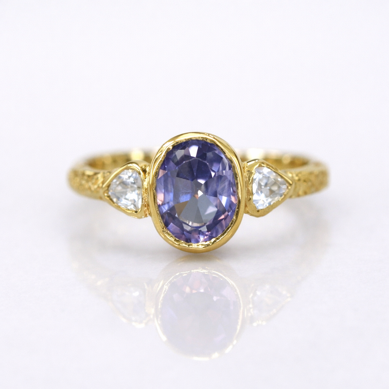 Custom gold ring featuring an oval lavender sapphire with trillion-cut diamond side stones, handcrafted by Alexis Pavlantos with a softly textured band.
