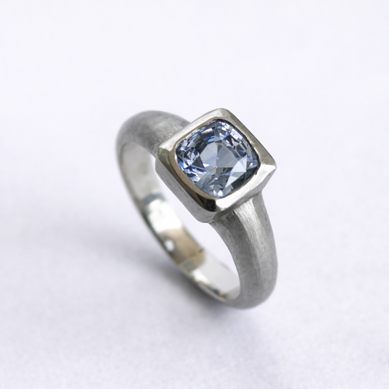Custom white gold solitaire ring featuring a cushion-cut blue sapphire, handcrafted with a softly contoured band by Albuquerque jeweler Alexis Pavlantos.